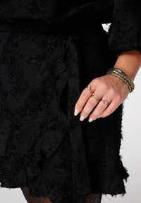 Zwarte getextureerde jurk met rafelige randen, aangesnoerd in de taille. Hand versierd met meerdere ringen en drie metalen armbanden. Lange, neutrale nagels.