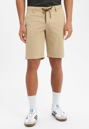 Beige knielange shorts met een bruine trekkoord, gedragen door een persoon met witte Adidas sneakers en witte enkelsokken.