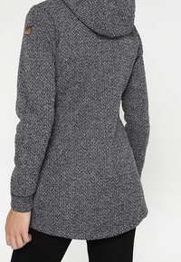 Grå tekstureret jakke med hætte, der har en tætsiddende talje, lange ærmer og et logo-patch på venstre skulder.