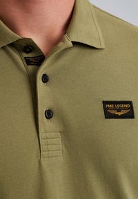 Olivi vihreä polo-paita, jossa on teksturoitu kangas, kolme mustaa nappia ja merkki, jossa lukee "PME Legend" mustalla ja kullalla.