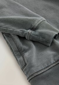Sweatshirt gris avec des poignets et un ourlet côtelés. Le tissu semble doux avec une texture usée. Les coutures sont visibles sur les manches et le corps.