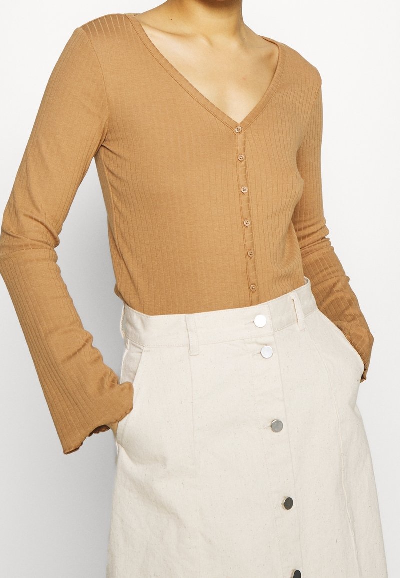 Haut à manches longues en côte, col en V, de couleur marron clair, avec une fermeture à boutons sur le devant et une silhouette cintrée, associé à une jupe beige.