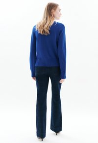 Blauwe gebreide trui met ribbels aan de mouwen en onderkant, gecombineerd met uitlopende donkere denim jeans. Model staat met de rug naar de camera tegen een witte achtergrond.