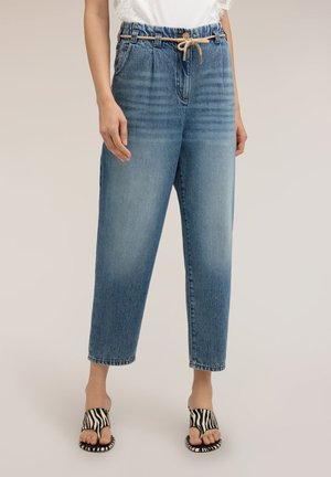 Donna che indossa jeans azzurri sbiaditi a vita alta con cintura beige in corda e sandali aperti con stampa a zebra, in piedi davanti a uno sfondo semplice.