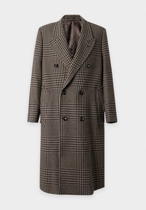 Man staat, draagt een lange geruite jas, crème knoophemd, grijze broek en zwarte schoenen, met één hand in de zak tegen een effen achtergrond.