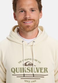 Sudadera de color crema claro con cordones, que presenta un logotipo gráfico verde y texto en la parte frontal. Tela suave y corte relajado.