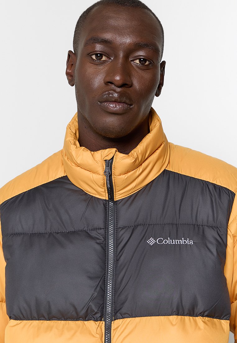Chaqueta tipo abrigo en colores amarillo y gris, con un cuello alto, cremallera frontal y el logo de Columbia en el pecho. Tejido texturizado con paneles.