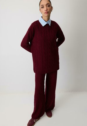 Kvinde står med armene bag ryggen iført en bordeaux kabelstrikket sweater og matchende vidde bukser over en lyseblå skjorte med krave.