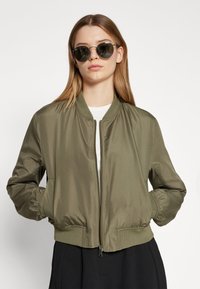 Olivově zelená bomber bunda vyrobená z hladkého materiálu, s předním zipem, elastickýmiManžetami a žebrovaným lemem, nošená přes bílé tričko.