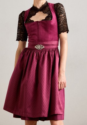 Femme portant une robe bordeaux texturée avec une ceinture à broche argentée sur un haut en dentelle noir à manches courtes et un col ras du cou, immobile.