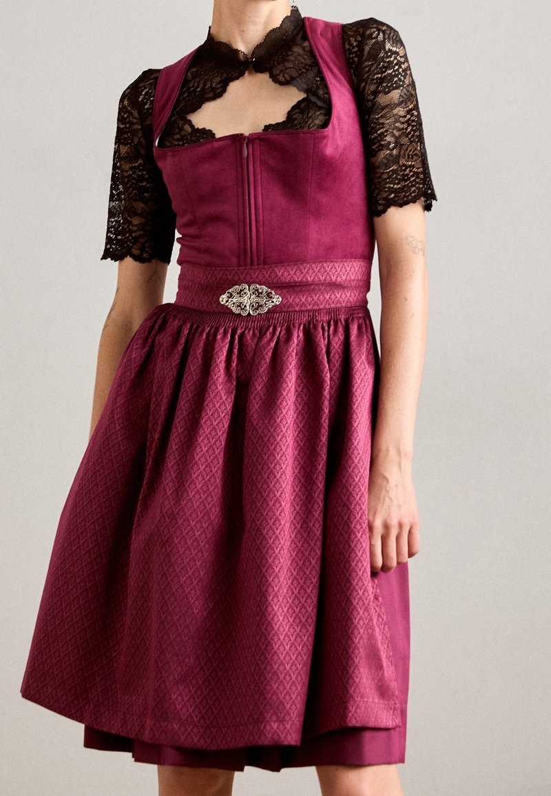 Femme portant une robe bordeaux texturée avec une ceinture à broche argentée sur un haut en dentelle noir à manches courtes et un col ras du cou, immobile.
