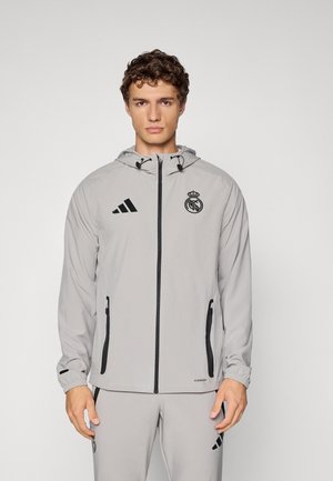 Fiatal férfi világosszürke Real Madrid melegítőben, cipzáras dzsekivel és kapucnival, egy sima világos háttér előtt áll.