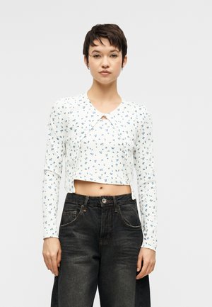 Kobieta o krótkich ciemnych włosach, ubrana w biały krótki kwiatowy top z długim rękawem i wysokim stanem przetartych czarnych dżinsów, stojąca na jednolitym tle.