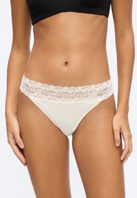 Frau trägt cremefarbene Bikini-Unterwäsche mit Spitzenbesatz und "Tommy Hilfiger"-Logo sowie ein schwarzes, kurzes Oberteil.