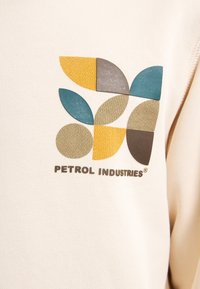 Tessuto color crema con un logo geometrico che presenta forme gialle, verdi, blu e marroni. Il testo recita "PETROL INDUSTRIES®."