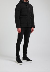 Czarna kurtka puchowa z kapturem, zapinana na zamek i guziki, z przednimi kieszeniami i pikowanym wykończeniem. Noszona z czarnymi dopasowanymi dżinsami i trampkami.