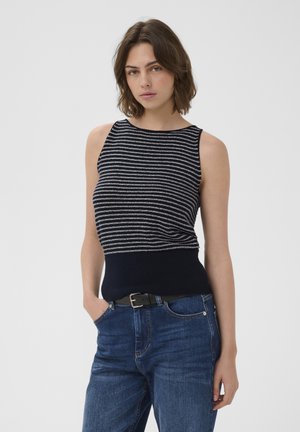 Frau mit schulterlangem braunem Haar, die ein ärmelloses marineblau-grau gestreiftes Oberteil und blaue Jeans mit schwarzem Gürtel trägt, steht.