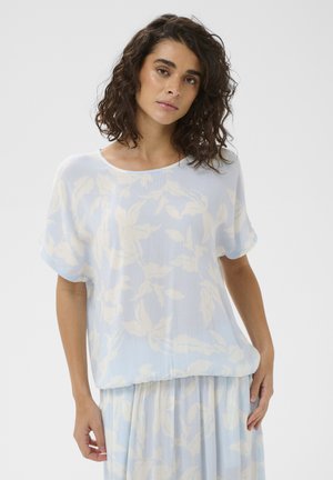 Kobieta o kręconych ciemnych włosach, ubrana w luźną, jasnoniebieską bluzkę w kwiaty z krótkim rękawem oraz pasującą spódnicę, na jednolitym tle.