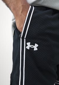 Pantalones cortos deportivos de malla negra con acentos blancos, bolsillos laterales y un logotipo de Under Armour. Presenta una textura transpirable y un diseño ajustado.