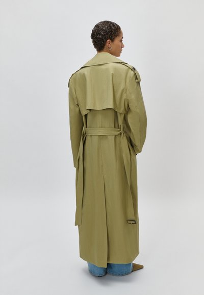 Άτομο με κοντά σγουρά μαλλιά που φοράει μακριά χακί καπαρντίνα, μπλε τζιν και μυτερά παπούτσια, γυρισμένος πλάτη σε απλό φόντο.