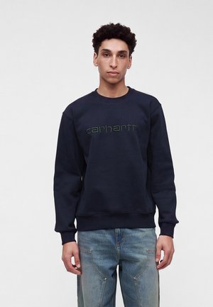 Joven con cabello rizado corto, vistiendo una sudadera Carhartt azul oscuro y vaqueros de mezclilla azul claro sobre un fondo claro y liso.