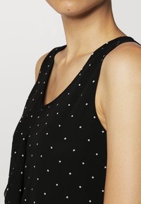 Top negro sin mangas con una textura suave, que presenta un patrón de pequeños lunares blancos. Escote redondo y corte relajado.