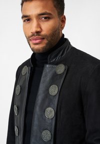 Schwarze Jacke aus Wildleder und Leder, mit hohem Kragen und sechs geprägten silbernen Knöpfen auf der Vorderseite, mit glatter Textur und tailliertem Schnitt.
