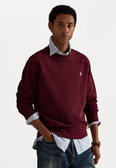 Burgundy-coloured huppari, jossa on pyöreä pääntie, kireät hihat ja brodeerattu logo; kerroksittain vaaleansinisellä raidallisella paidalla, jonka hihat ovat näkyvissä.