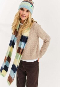 Beżowy sweter dziergany w splot warkoczowy z okrągłym dekoltem, uzupełniony wielokolorowym, kratkowanym szalikiem oraz miękką, niebieską czapką z dzianiny z pomponem.