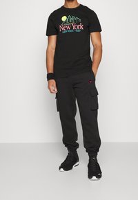 Svart t-shirt med färggrafik "New York"; kombinerad med svarta cargojoggers. Modellen bär svarta träningstävlar med vita detaljer.