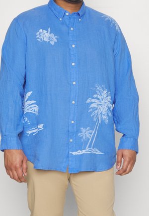 Homme portant une chemise bleue à manches longues avec des imprimés tropicaux blancs et un pantalon beige sur un fond uni.