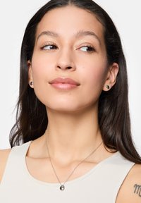 Zilveren ketting met een druppelvormige hanger; model draagt kleine schouderoorbellen; gladde huid; lichte mouwloze top; neutrale achtergrond.