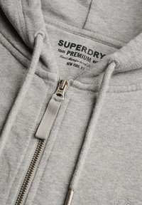 Zbliżenie na szarą bluzę z kapturem zapinaną na suwak z metalowym suwakiem oraz czarnym nadrukowanym napisem marki „Superdry Premium New York, NY” wewnątrz kołnierza.
