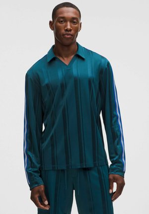 Man draagt een teal gestreept shirt met lange mouwen met blauwe en witte strepen op de mouwen en bijpassende broek tegen een effen achtergrond.