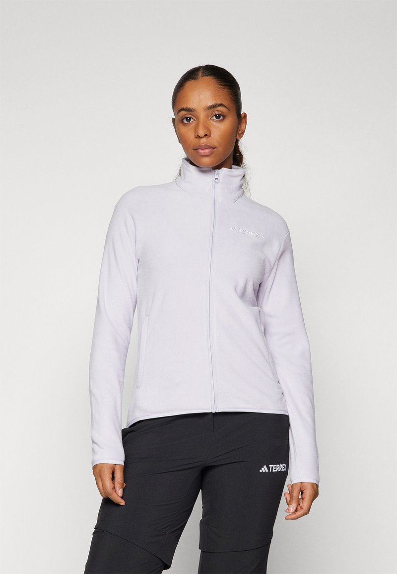 Helllila Fleecejacke mit hohem Kragen, Frontreißverschluss und gesticktem Logo. Kombiniert mit schwarzen Hosen, die ein Logo auf dem Oberschenkel haben.