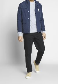 Marineblaues Jacket mit mehreren Taschen, grauer Pullover, schwarze Hose und hellgelbe Sneaker. Glatter Stoff mit tailliertem Schnitt.