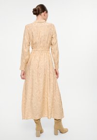 Robe beige à manches longues avec un motif de vagues texturé, taille ajustée et fentes latérales, assortie à des bottes montantes beige.