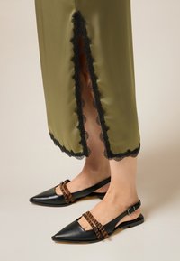 Scarpe slingback in pelle nera con punta appuntita, dotate di due cinturini a strisce marroni. Gonna verde oliva con bordo in pizzo nero all'orlo.