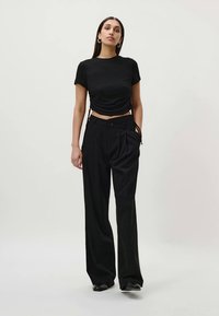 Kvinne iført svart rynket kortermet crop topp, høyt liv, vide svarte bukser og svarte sko, stående mot hvit bakgrunn.