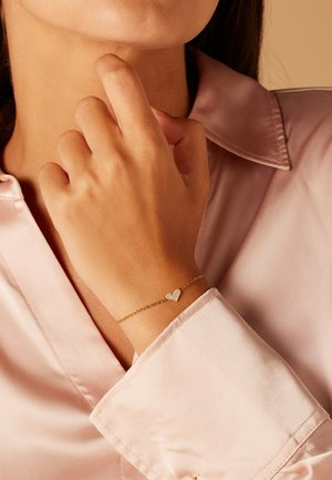Femme portant un délicat bracelet chaîne en or avec un pendentif en forme de cœur blanc, vêtue d'un chemisier en satin rose clair, main près du menton.