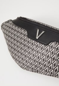 Svart och grå väska med geometriskt mönster, med en slät läderdetalj och silvrig dragkedja, med en framträdande metallisk "V"-logotyp.