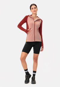 Aktivjacke in Pink und Maroon mit einem Reißverschluss vorne, langen Ärmeln und Taschen. Kombiniert mit schwarzen Shorts und Sneakers mit orangefarbenen Akzenten.