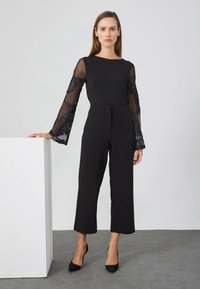 Mulher vestindo um macacão preto com mangas de renda floral transparente e saltos pretos, de pé com uma das mãos apoiada em um bloco branco.
