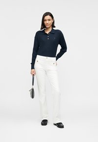 Μπλε ναυτικού πλεκτό πόλο με μακριά μανίκια, λευκό γιακά, χρυσά κουμπιά, σε συνδυασμό με λευκό παντελόνι με ψηλή μέση και μαύρα παπούτσια τύπου slip-on.