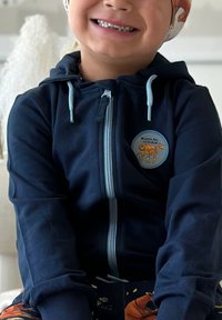 Tummansininen vetoketjullinen huppari, joka on valmistettu pehmeästä kankaasta. Hupparissa on vaaleansinisiä yksityiskohtia ja pyöreä kangasmerkki, jossa on hevosen grafiikka.