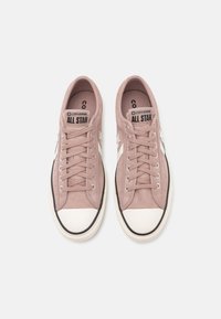 Baskets en daim rose avec capuchon en caoutchouc blanc, semelle extérieure noire et six œillets pour les lacets. Logo de la marque sur la languette : "Converse All Star."