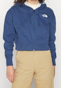 Felpa blu tagliata con zip frontale, due tasche, cappuccio con coulisse e logo bianco sul lato sinistro del petto. Tessuto misto cotone, polsini a costine.