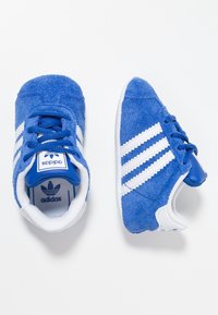 Modré semisové tenisky pro batolata s bílými pruhy a modrými detaily. Mají polstrovaný límec a modrý jazyk s logem. Texturovaná gumová podrážka.