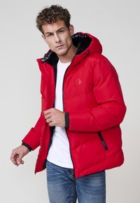 Chaqueta aislante roja con capucha y puños forrados de piel sintética negra. Presenta un logo en el pecho y bolsillos laterales con cremallera. Textura exterior suave.