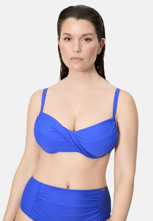 Mujer con el cabello largo y mojado, que lleva un bikini azul brillante con un top estilo cruzado y pantalones de talle alto, sobre un fondo liso.